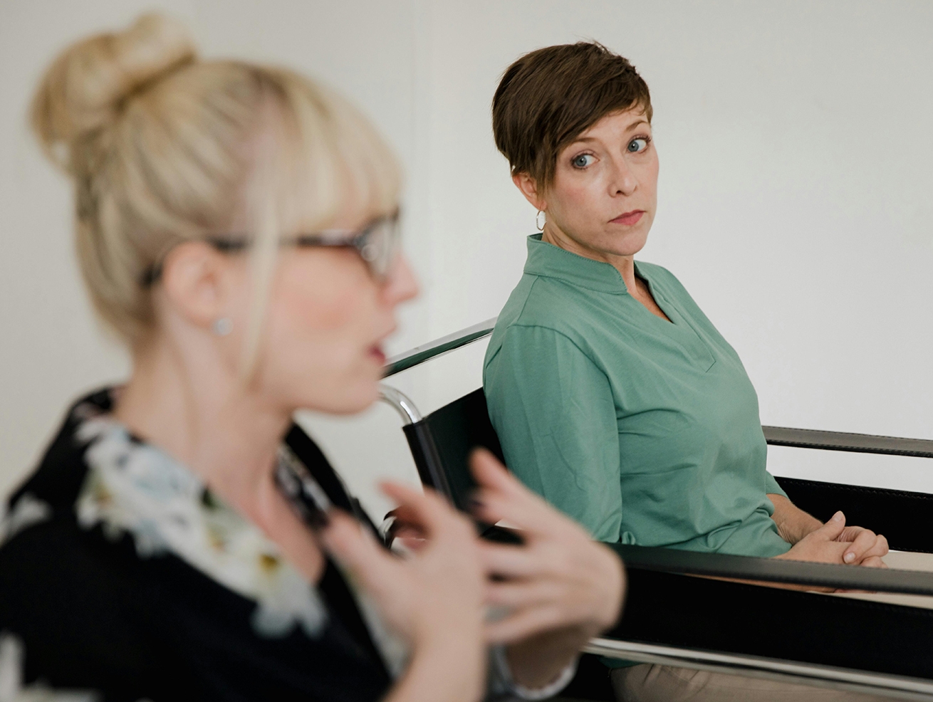 A blonde woman with glasses and a brunette woman in a green shirt discussing their business partnership dispute in a counsellor’s office.